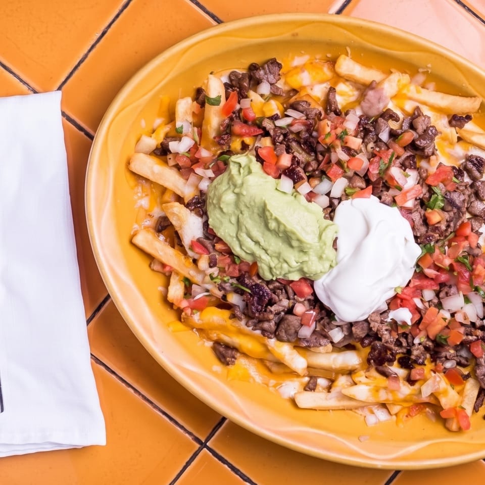 Loaded cheesy fries topped with diced steak, black beans, pico de gallo, guacamole, and sour cream on a colorful tiled table.