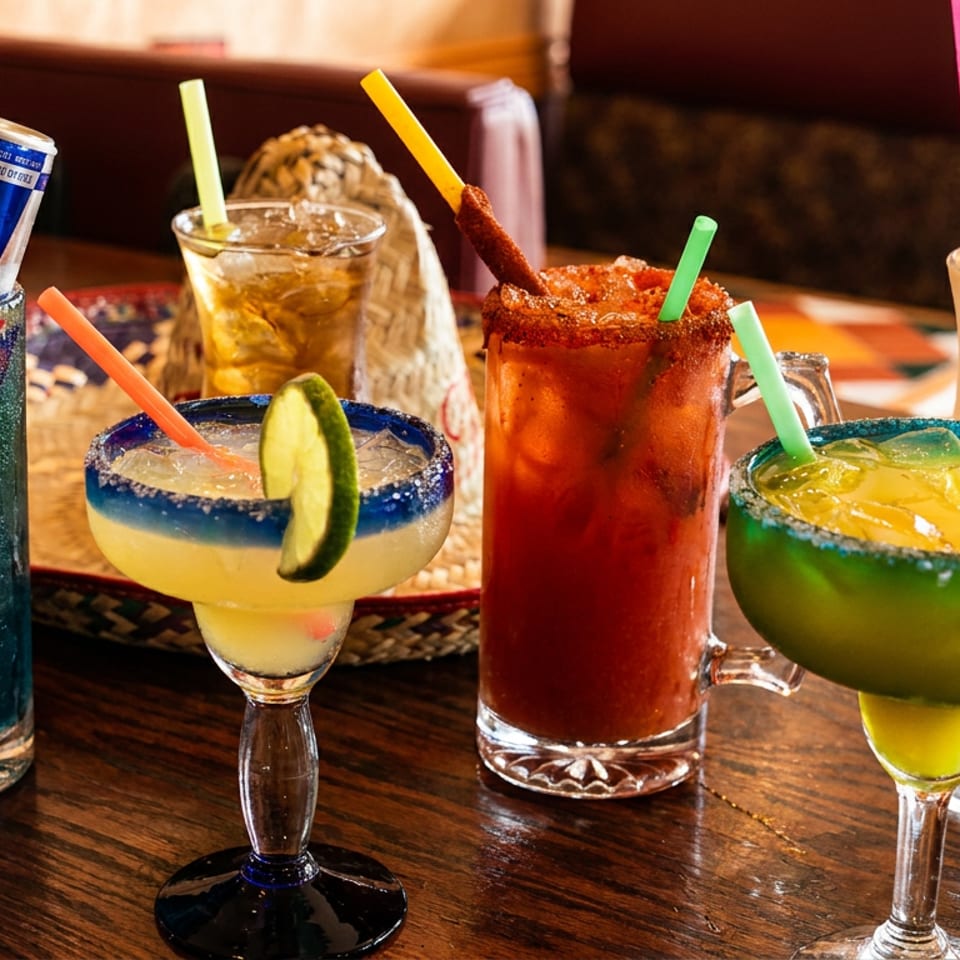 A variety of colorful drinks, including margaritas, a Red Bull cocktail, and a milkshake, on a wooden table.