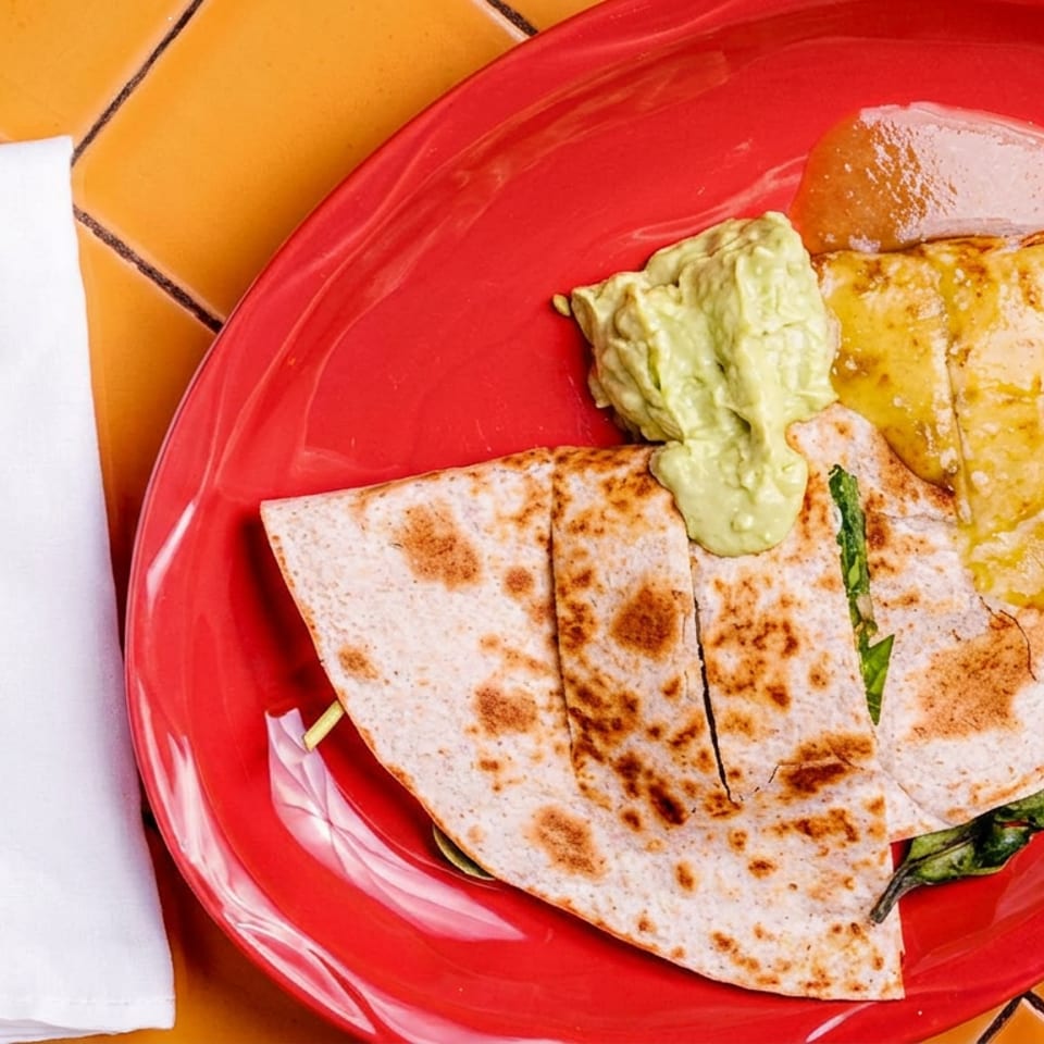 Quesadilla slices with guacamole and salsa verde on a red plate next to a fork and napkin.